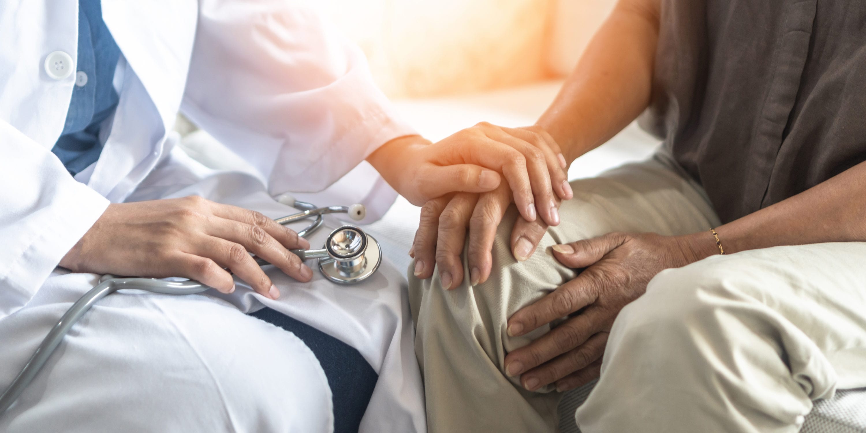 Läkare sitter ned med patient och håller handen på hens hand. I andra handen håller läkaren i ett stetoskop.